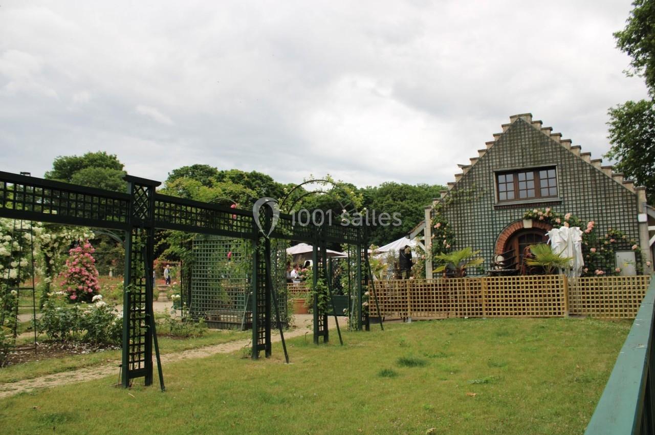 Jardin avec pergolas en métal, pelouse, rosiers en fleurs et bâtiment en briques avec toit à pignon en arrière-plan.