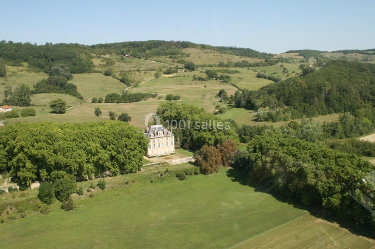 Vue aérienne d'un château entouré d'arbres et de prairies verdoyantes dans un paysage vallonné.