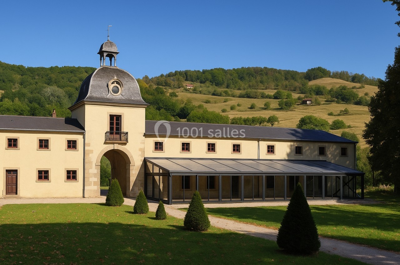 Bâtiment ancien avec clocher, grandes fenêtres et verrière, entouré de pelouses et collines verdoyantes.