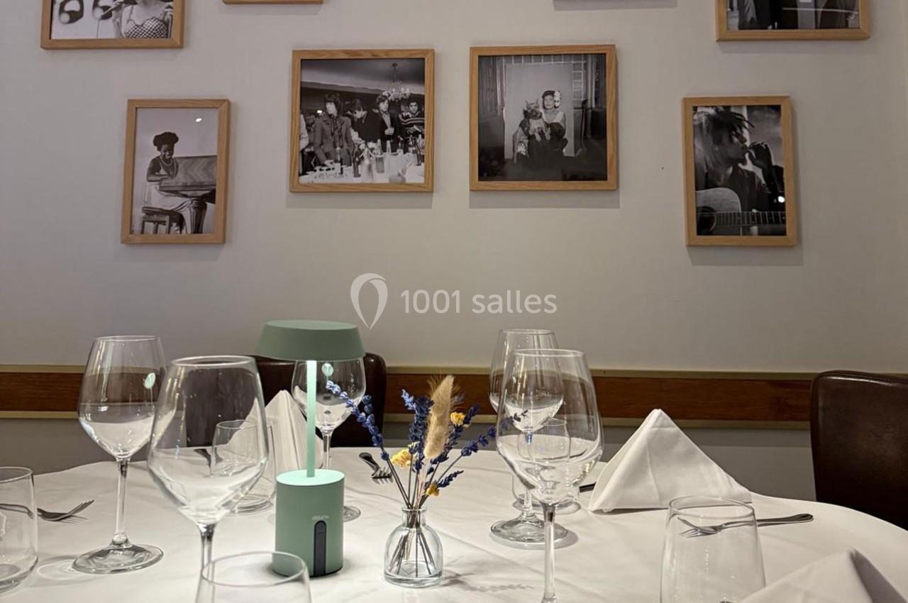 Table dressée avec des verres, serviettes pliées et un petit vase au centre, devant un mur décoré de photos encadrées.