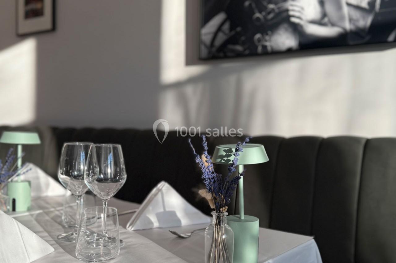 Table dressée avec nappes blanches, verres à vin, serviettes pliées et décoration florale dans un restaurant lumineux.