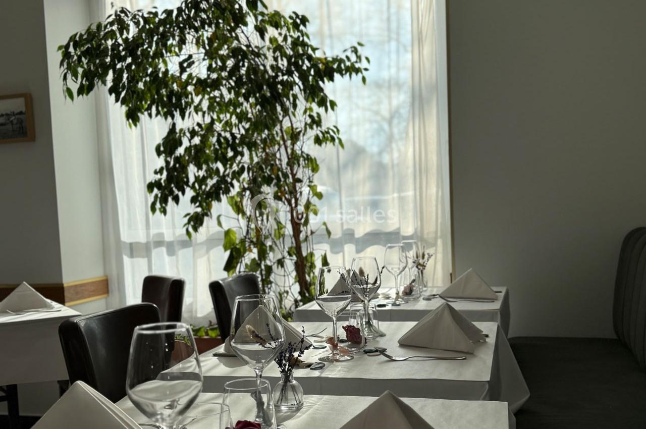 Salle de restaurant lumineuse avec tables dressées, nappes blanches, verres et serviettes pliées, près d'une grande plante.
