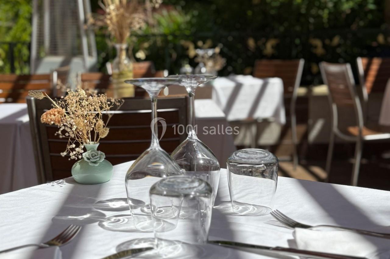 Table dressée en extérieur avec nappes blanches, verres, couverts et décoration florale sous une lumière ensoleillée.