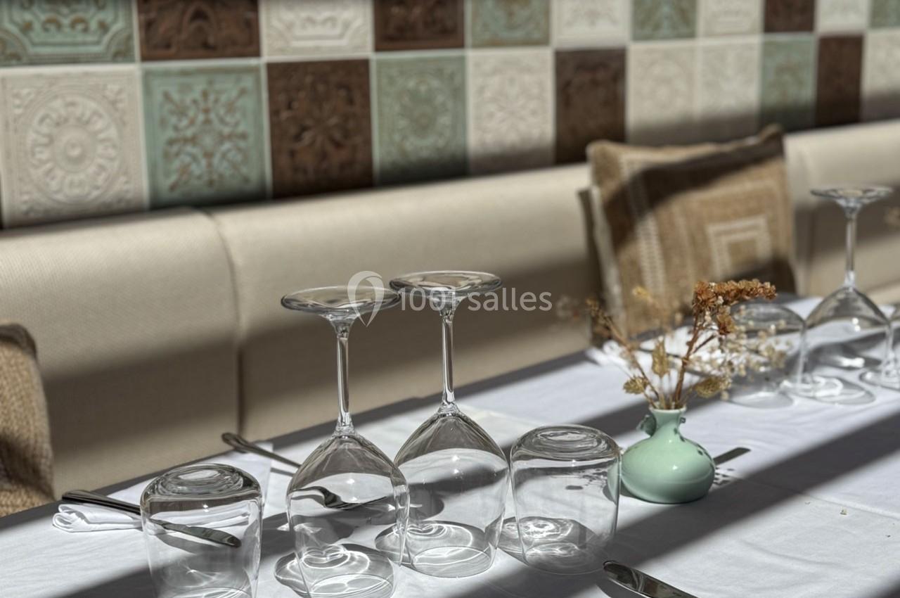 Verres retournés et vase avec fleurs séchées sur une table dressée, devant un mur de carreaux décoratifs.