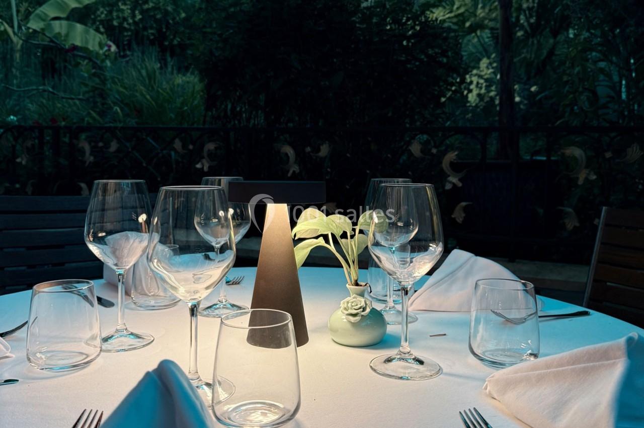 Table dressée en extérieur avec des verres, une lampe centrale et un petit vase contenant une plante, ambiance nocturne.