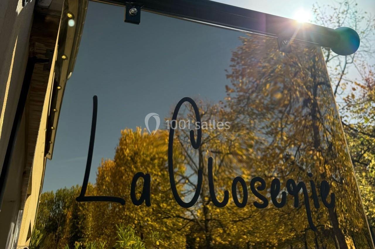Panneau en verre avec l'inscription ’La Closerie’, reflétant des arbres aux feuilles dorées sous un ciel ensoleillé.