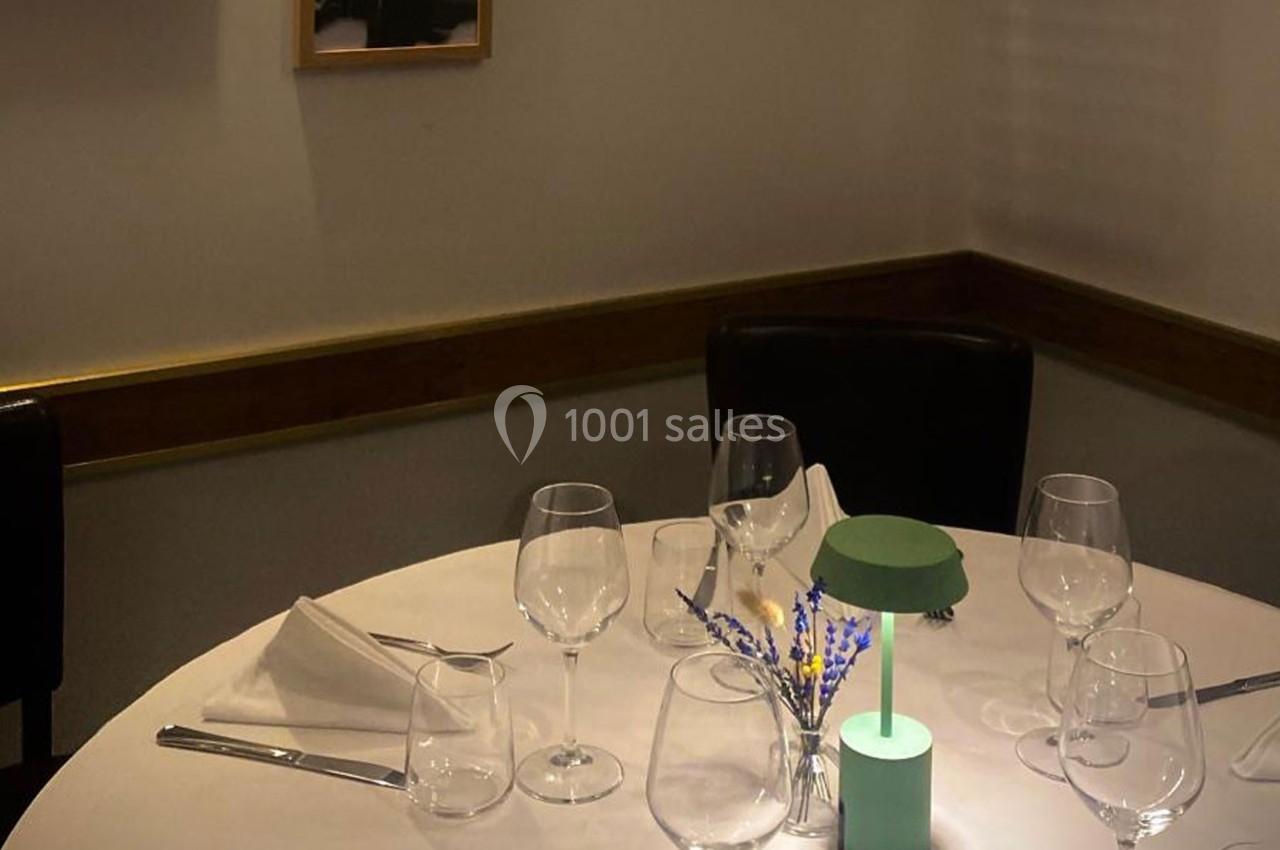 Table dressée avec nappe blanche, verres à vin, serviettes pliées, petite lampe verte et décoration florale.