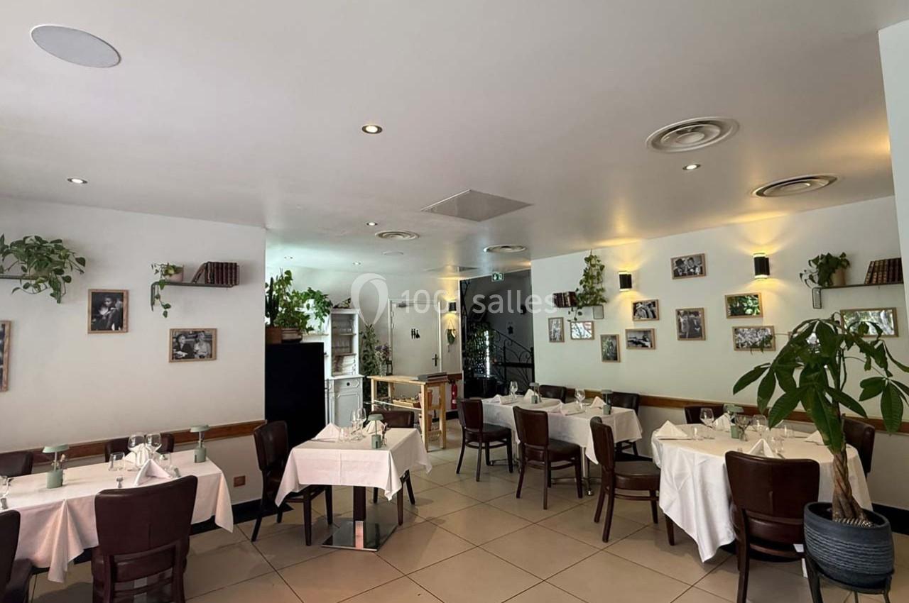 Salle de restaurant lumineuse avec tables dressées, chaises en bois, plantes décoratives et photos accrochées aux murs.