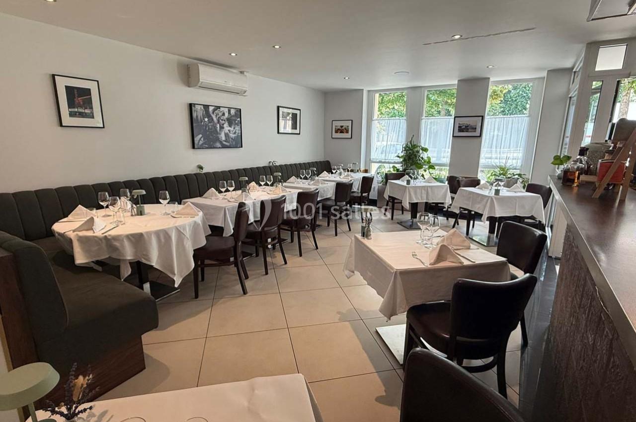 Salle de restaurant lumineuse avec tables dressées, nappes blanches et grandes fenêtres donnant sur l'extérieur.