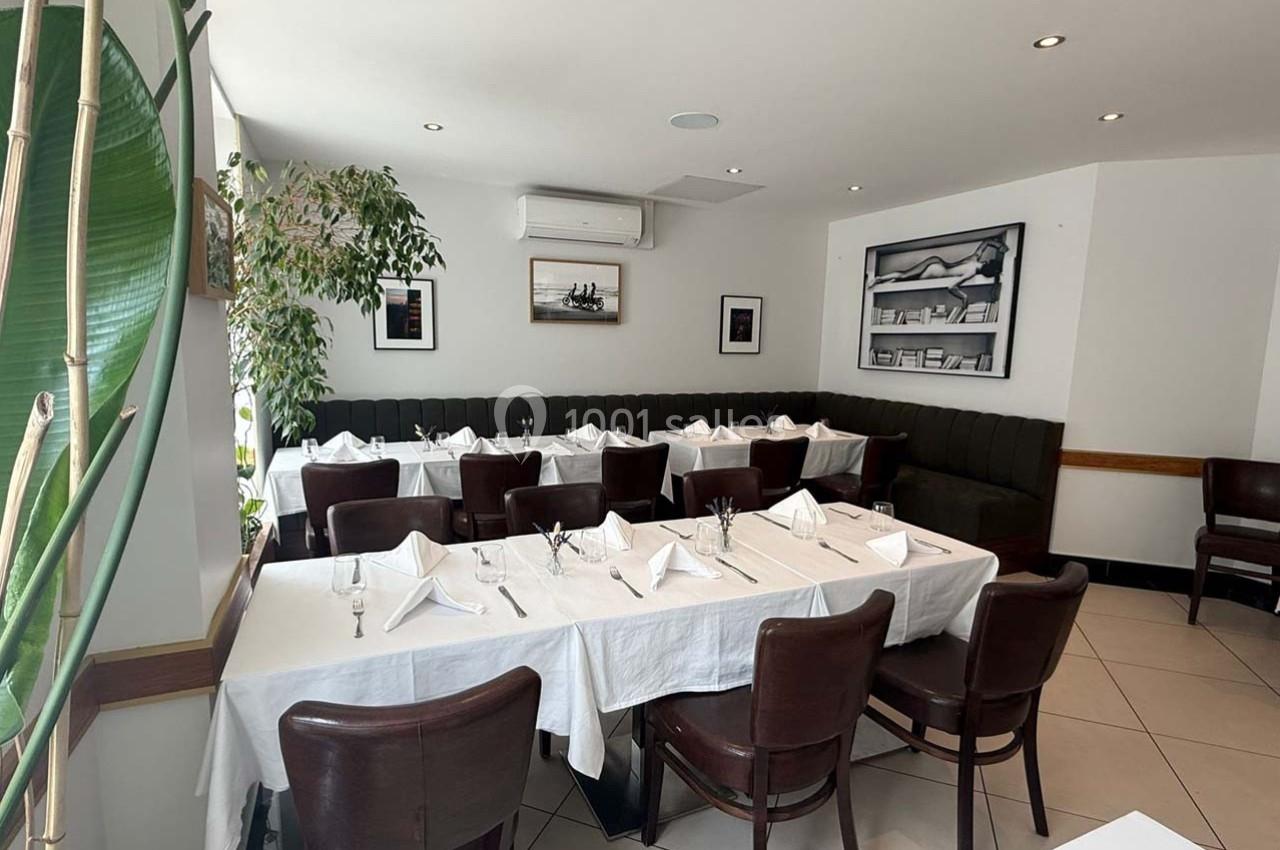 Salle de restaurant lumineuse avec tables dressées de nappes blanches, chaises en bois et plantes décoratives.