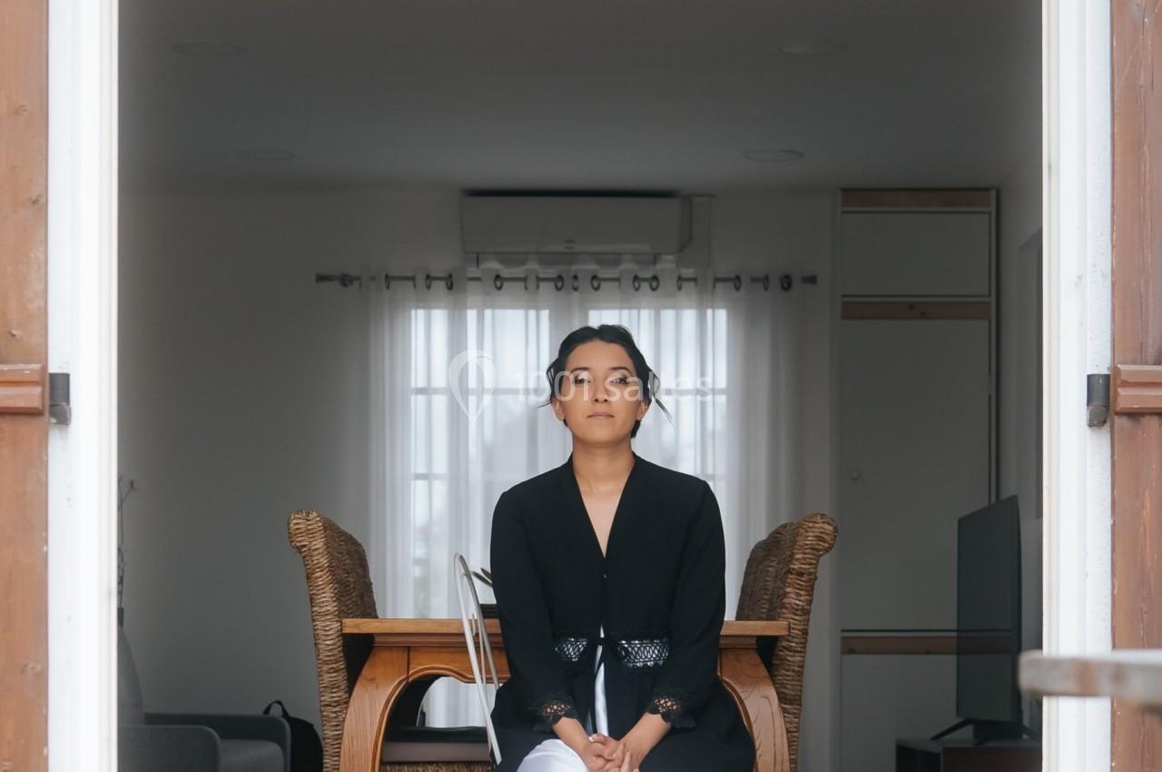 Femme assise sur une chaise dans l'encadrement d'une porte ouverte, intérieur lumineux avec table et chaises en arrière-plan.