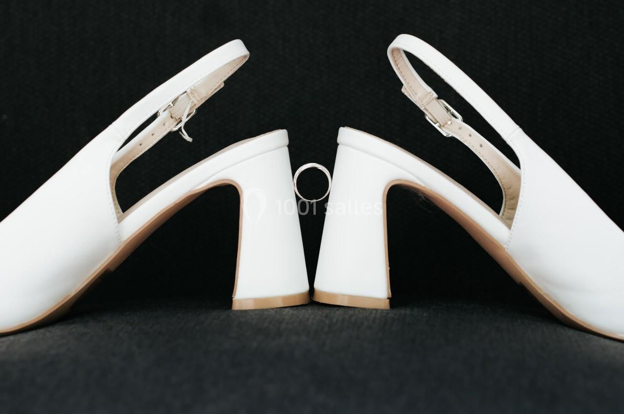 Deux chaussures blanches à talons hauts disposées face à face avec une bague suspendue entre elles.