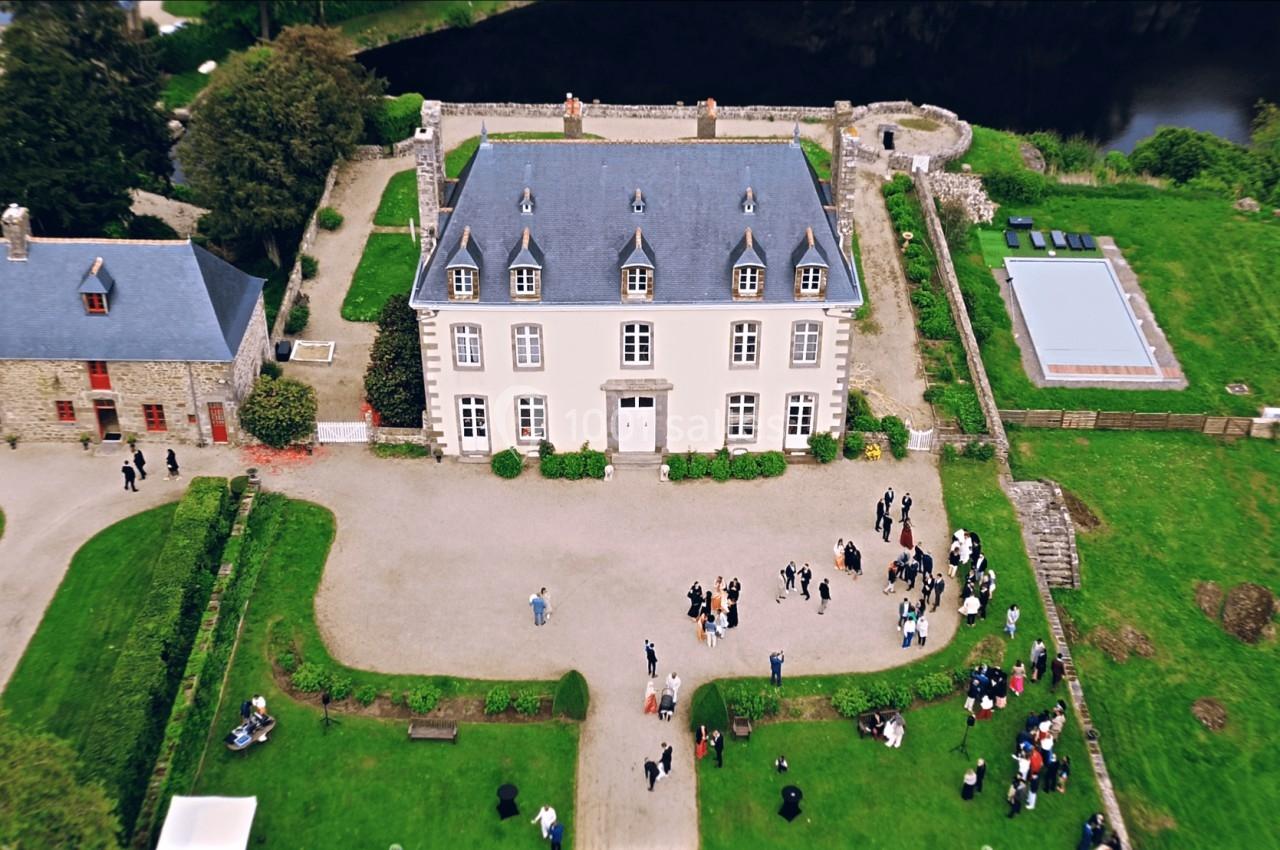 Vue aérienne d'un manoir entouré de pelouses, avec des invités rassemblés devant et une aire de jeux colorée à gauche.