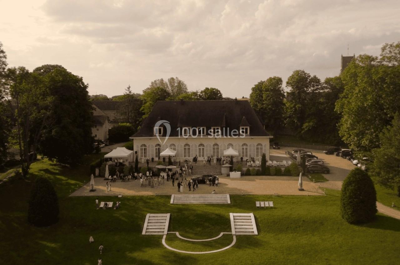 Vue aérienne d'un manoir entouré de verdure, avec des invités rassemblés sur une terrasse et une pelouse aménagée.