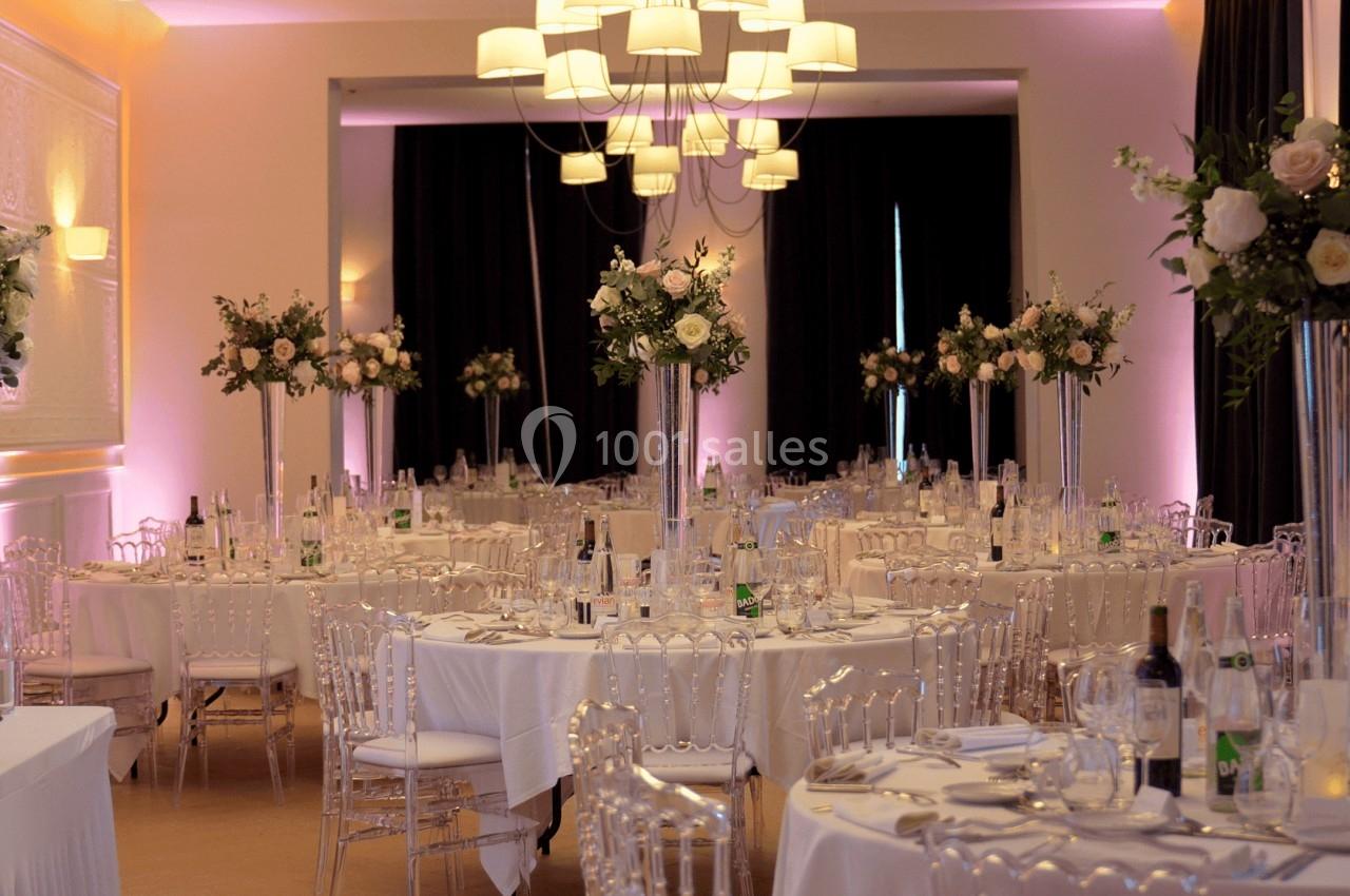 Salle de réception élégante avec tables dressées, chaises transparentes et décorations florales sur des vases hauts.