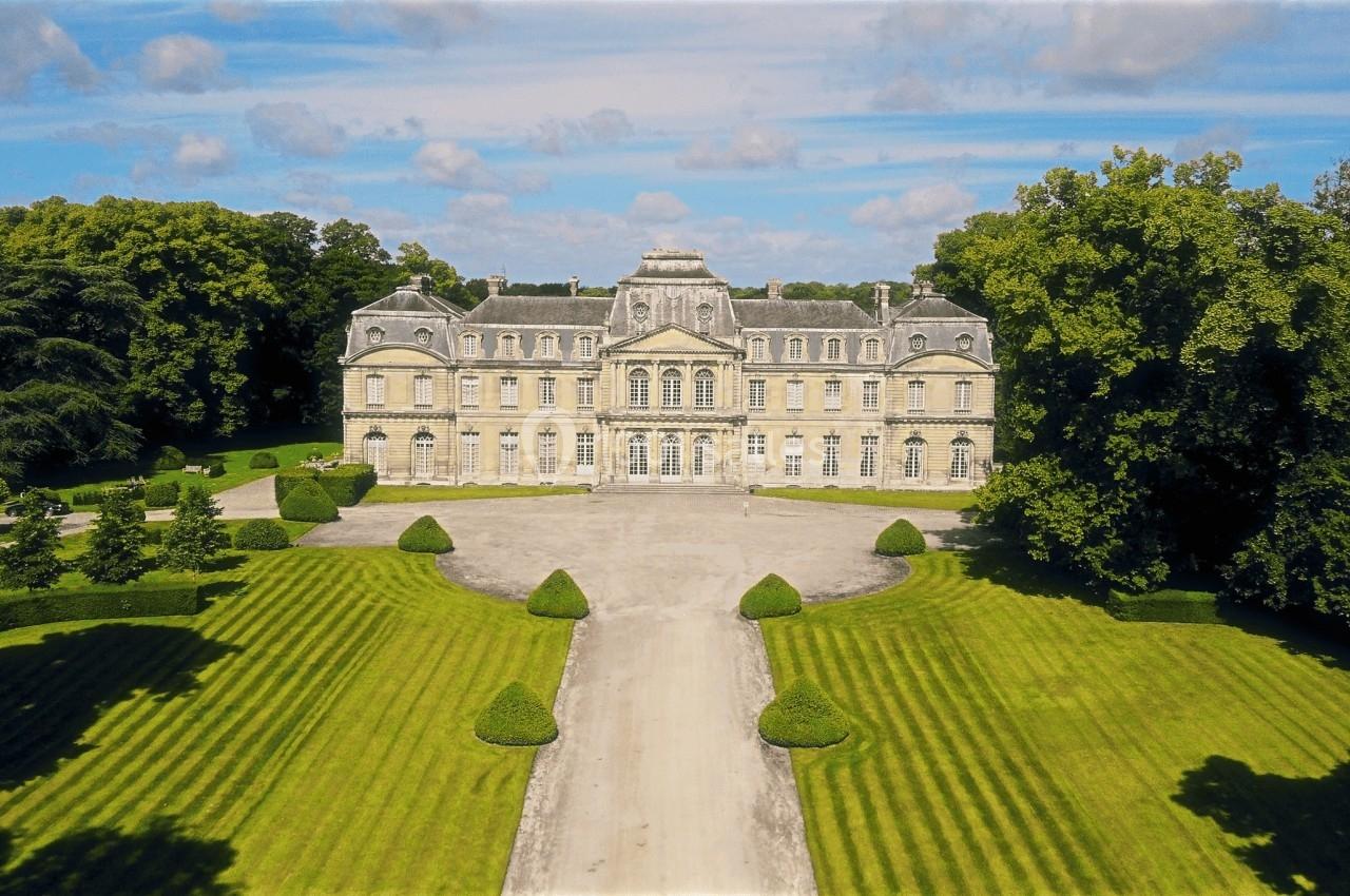 Façade d'un château entouré de jardins symétriques et d'arbres sous un ciel partiellement nuageux.