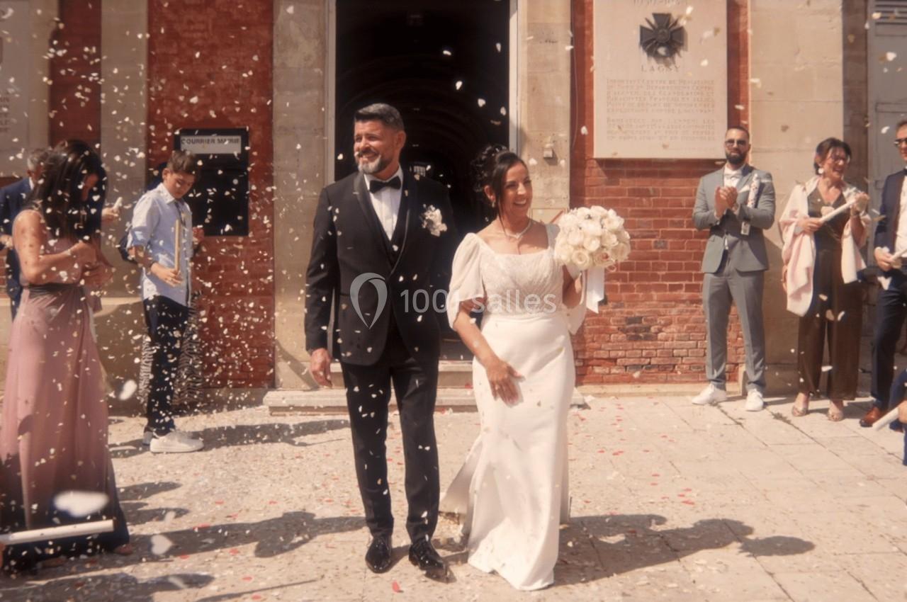 Un couple en tenue de mariage sort d'un bâtiment sous une pluie de confettis, entouré d'invités applaudissant.