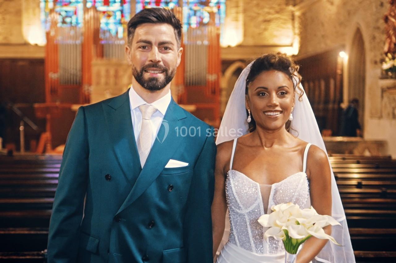 Un couple en tenue de mariage pose dans une église, avec des bancs en bois et un vitrail coloré en arrière-plan.