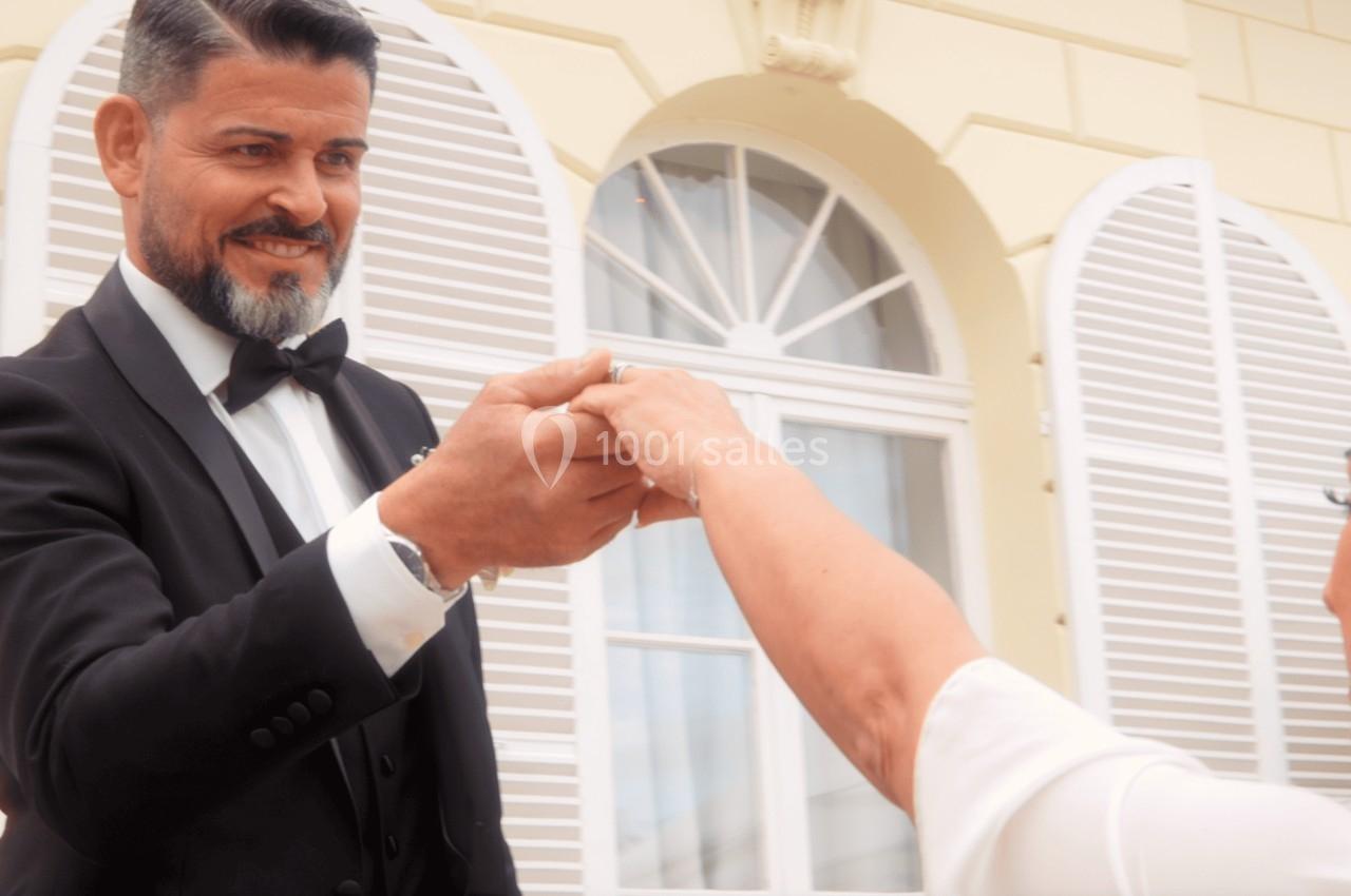 Un homme en costume noir tient la main d'une femme en robe blanche devant un bâtiment aux volets blancs.