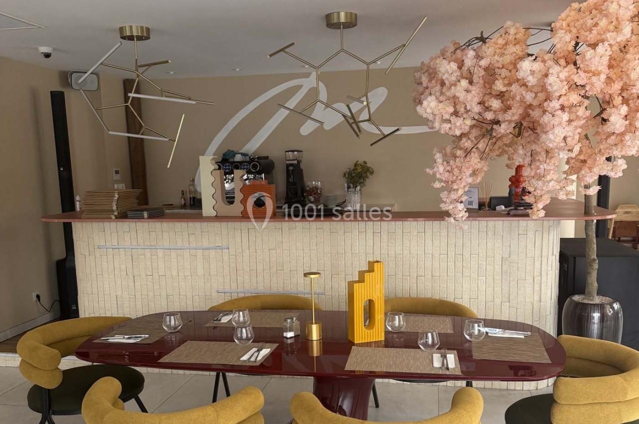 Salle de restaurant avec table dressée, chaises jaunes, comptoir en arrière-plan et décoration florale rose.