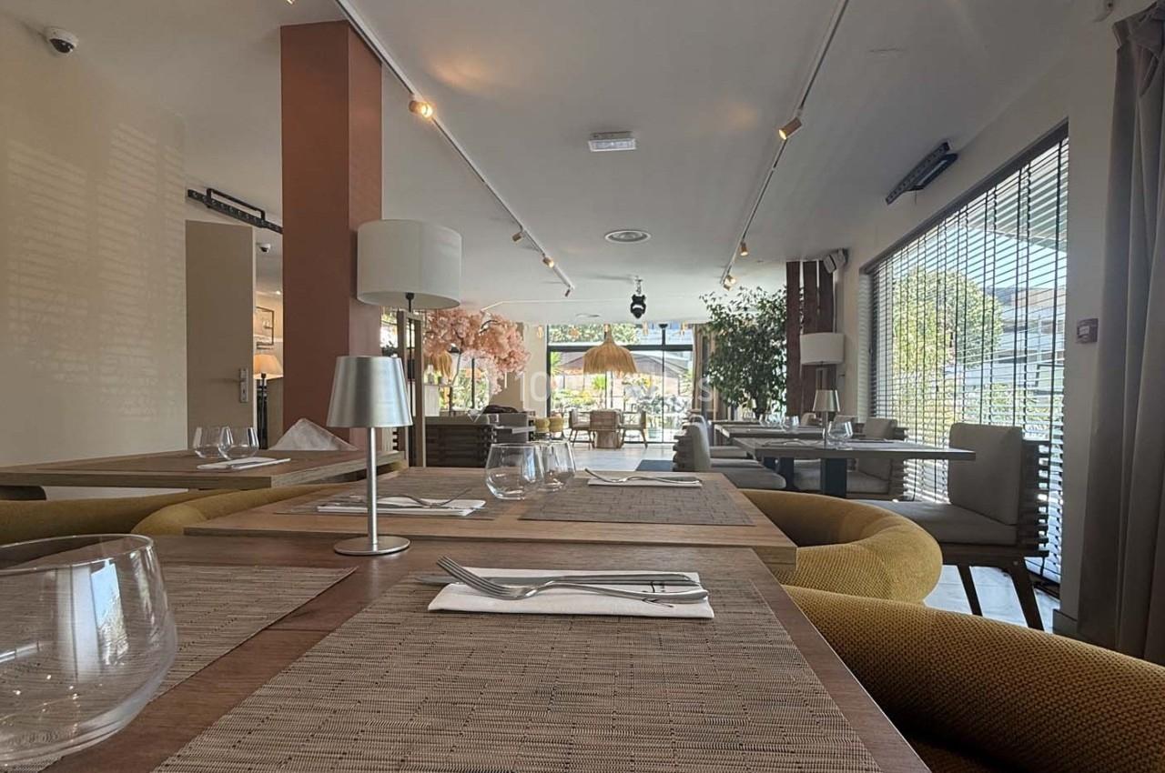 Salle de restaurant lumineuse avec tables dressées, chaises confortables et grandes baies vitrées laissant entrer la lumière…