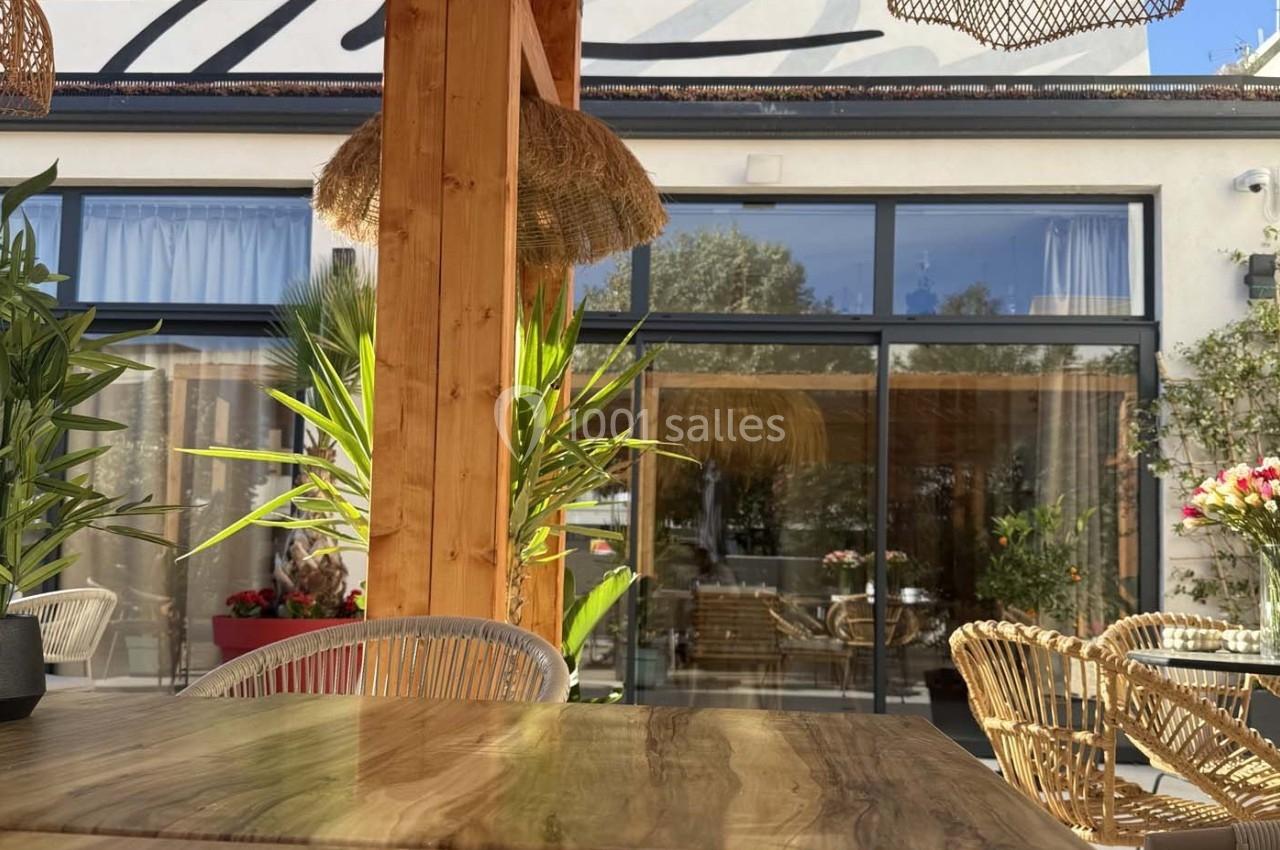 Terrasse ensoleillée avec table en bois, chaises en rotin, plantes et vue sur une baie vitrée donnant sur l'intérieur.