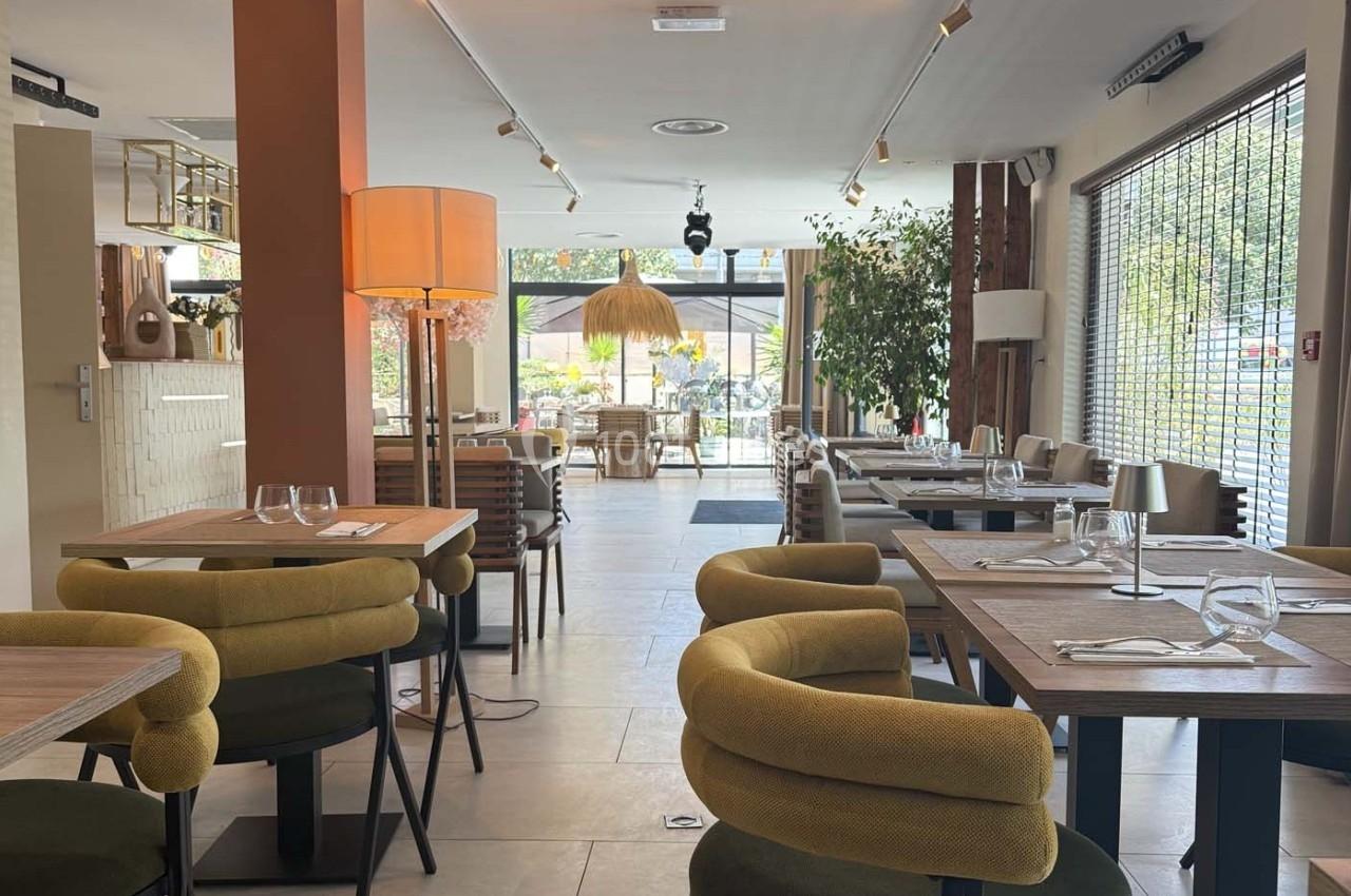Salle de restaurant lumineuse avec tables en bois, chaises rembourrées et vue sur une terrasse extérieure arborée.