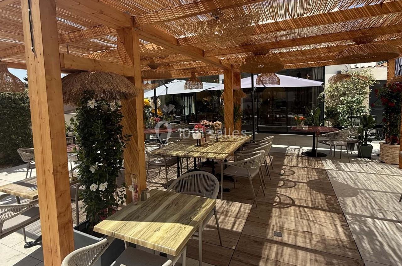 Terrasse en bois avec tables, chaises et pergola, entourée de plantes et éclairée par la lumière naturelle.