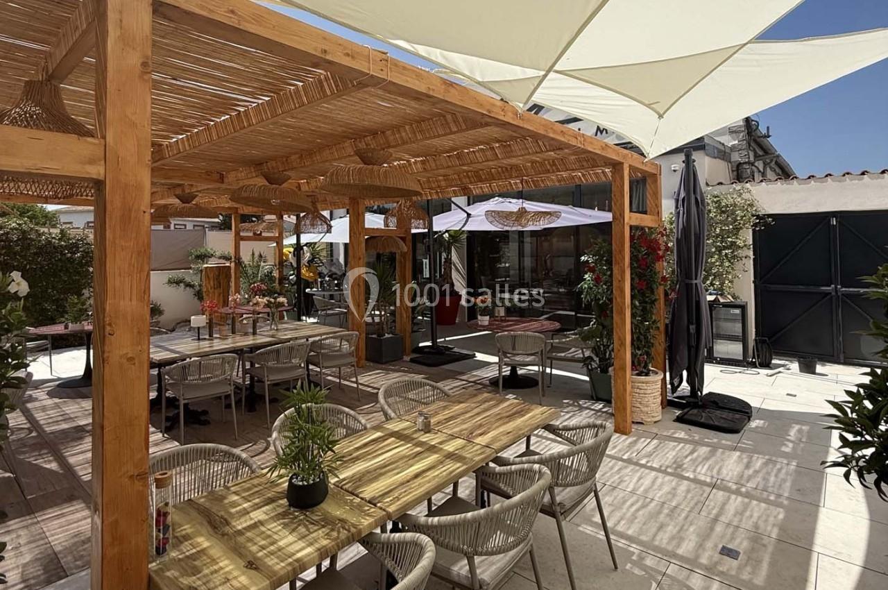 Terrasse extérieure avec pergola en bois, tables et chaises, entourée de plantes et protégée par des parasols.