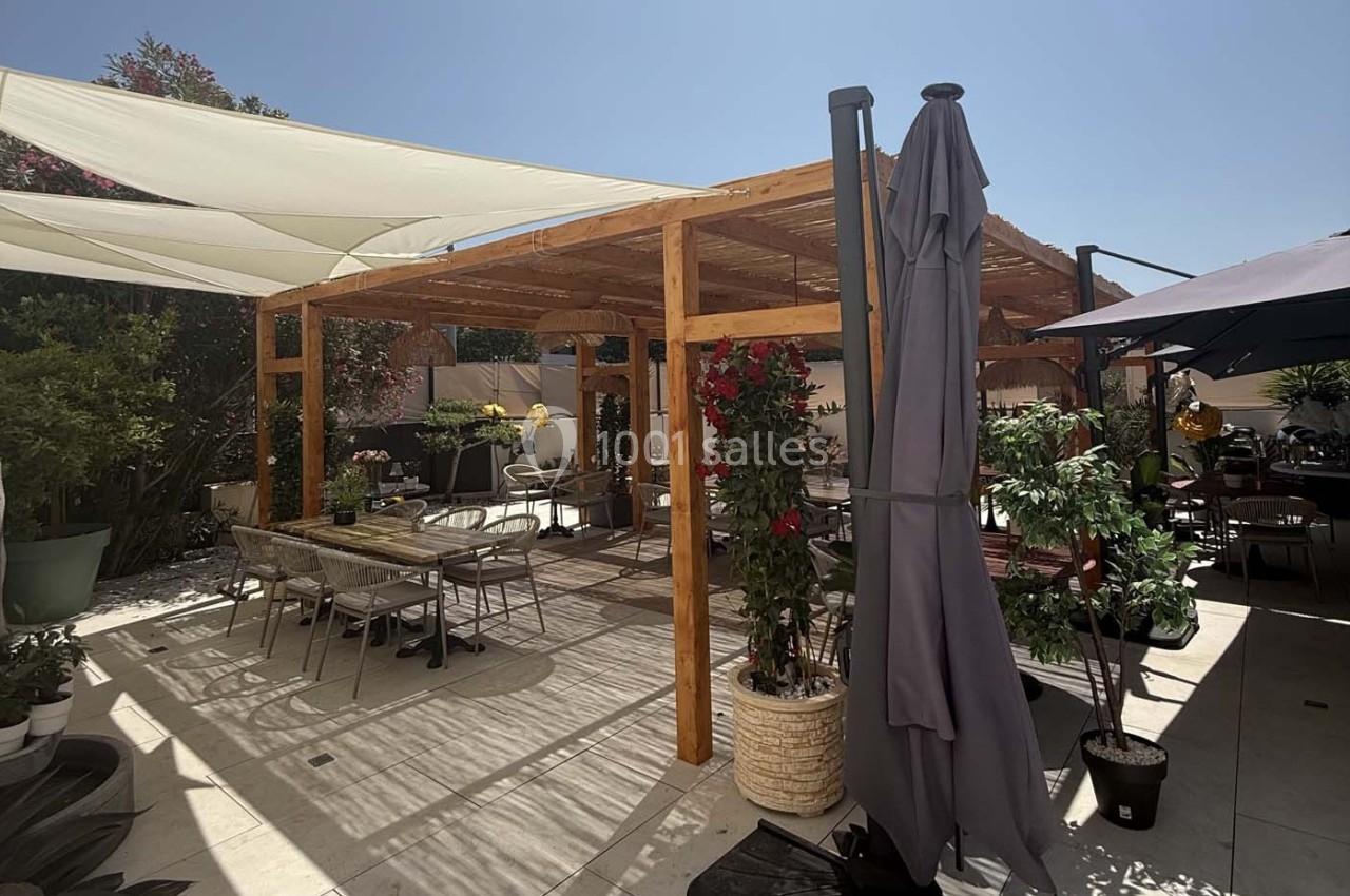 Pergola en bois avec tables et chaises sur une terrasse ensoleillée, entourée de plantes et de parasols.