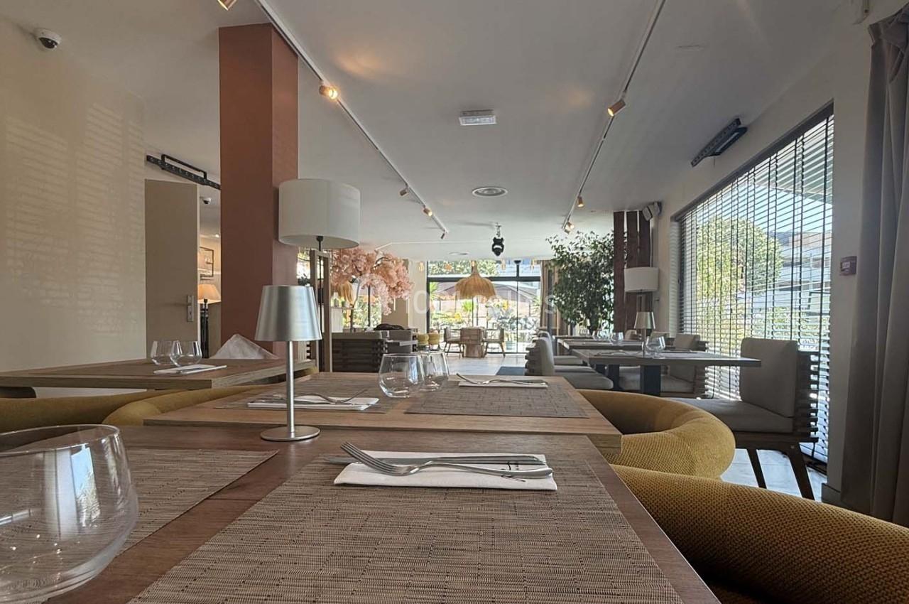 Salle de restaurant lumineuse avec tables dressées, chaises confortables et grandes baies vitrées donnant sur l'extérieur.