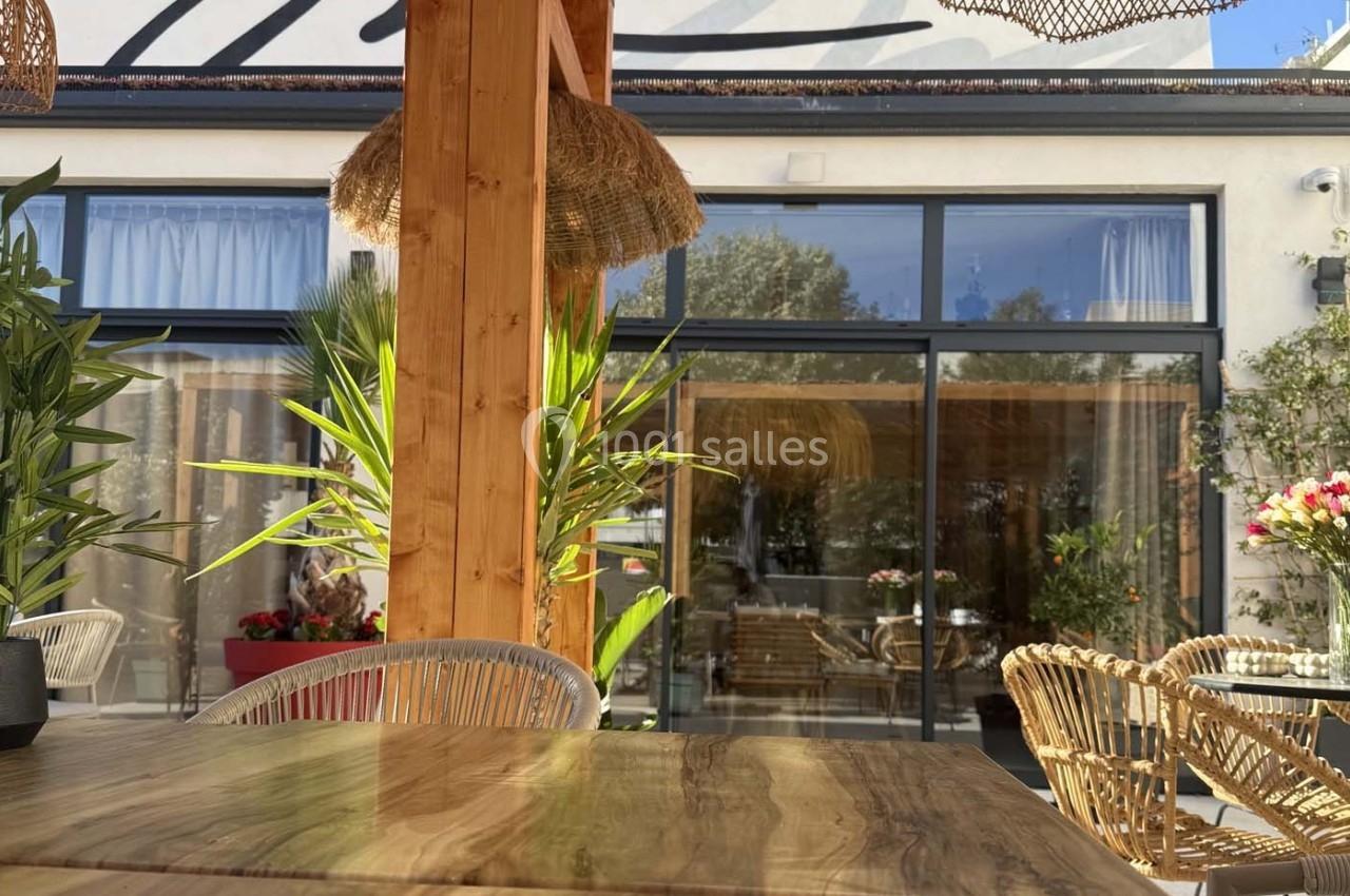 Terrasse ensoleillée avec table en bois, chaises en rotin et plantes, devant une baie vitrée donnant sur un intérieur…