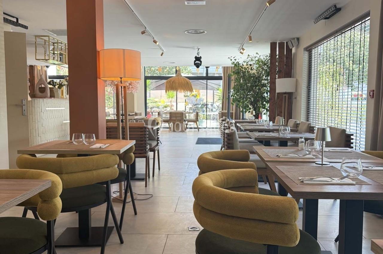 Salle de restaurant lumineuse avec tables dressées, chaises rembourrées et grandes baies vitrées donnant sur une terrasse.