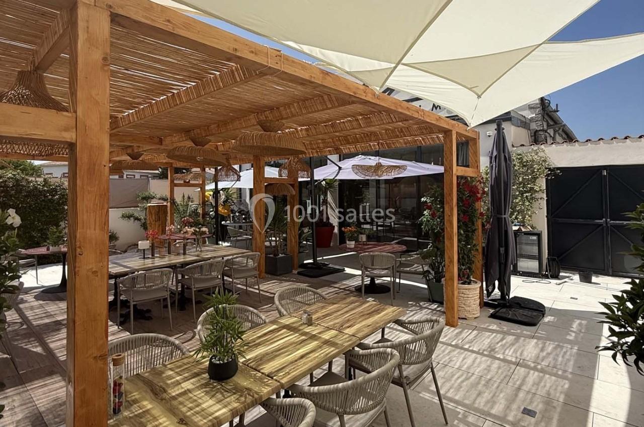 Terrasse extérieure avec pergola en bois, tables, chaises et plantes, ombragée par des voiles tendues.