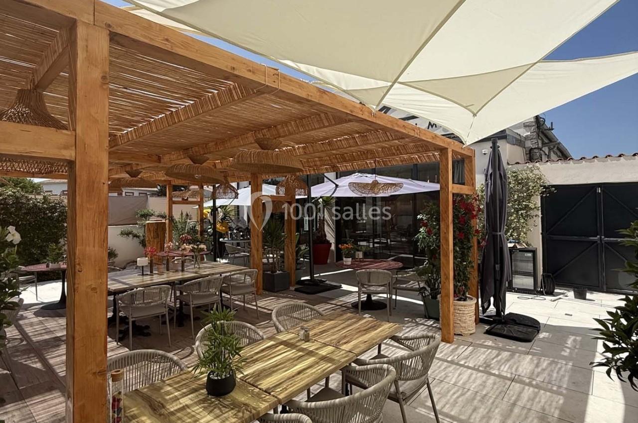 Terrasse extérieure avec pergola en bois, tables et chaises, entourée de plantes et protégée par des parasols blancs.