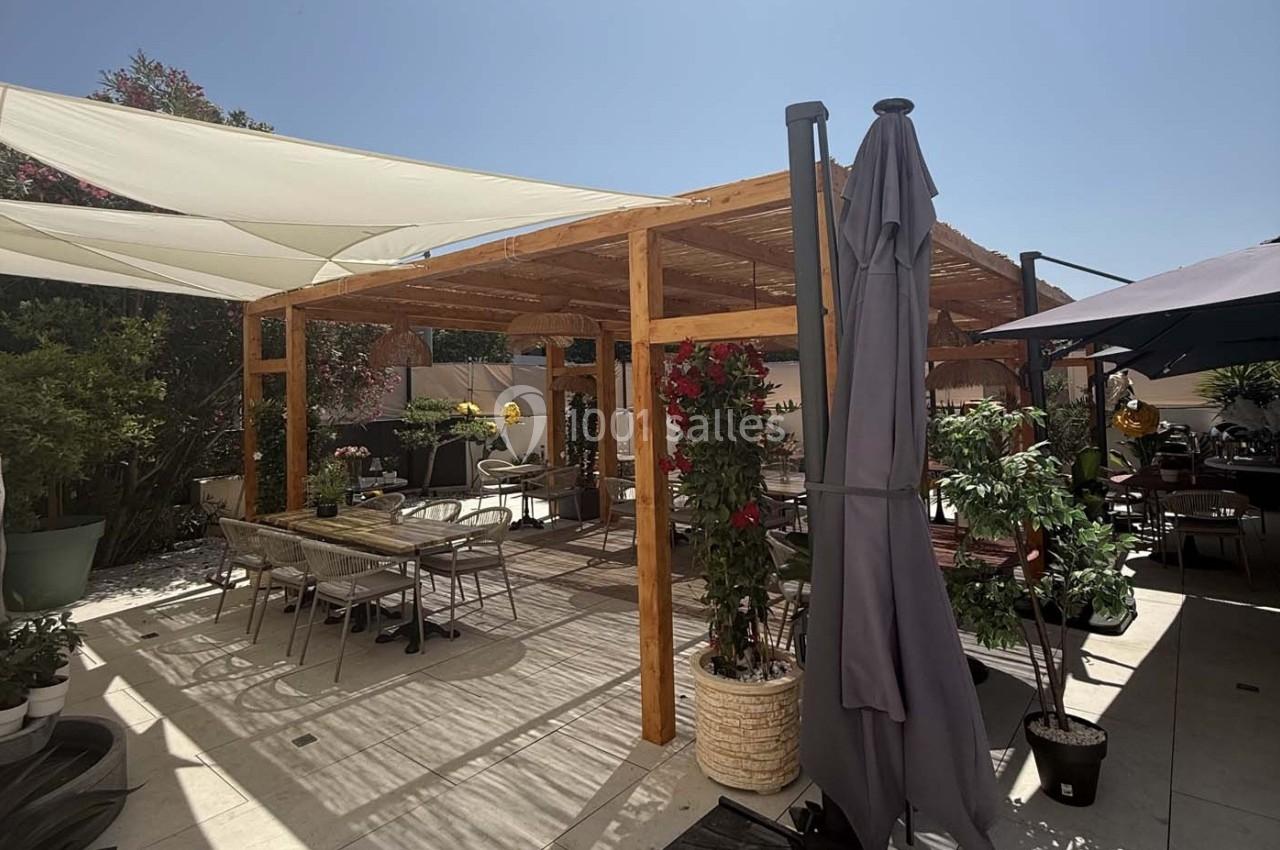 Pergola en bois avec voiles d'ombrage, entourée de plantes et de tables installées sur une terrasse ensoleillée.