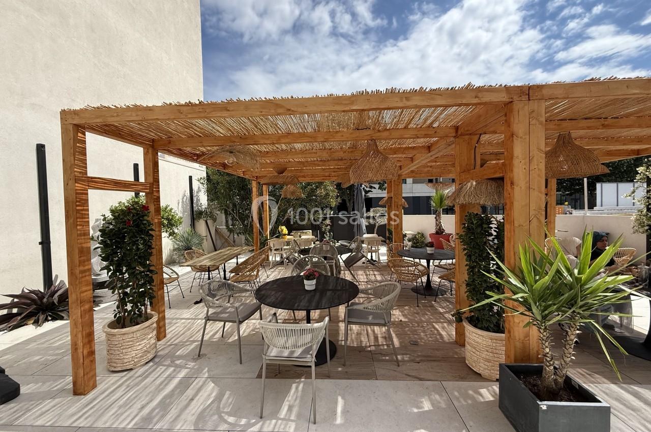 Pergola en bois avec toit en canisses, entourée de plantes en pots, mobilier de jardin et ciel partiellement nuageux.