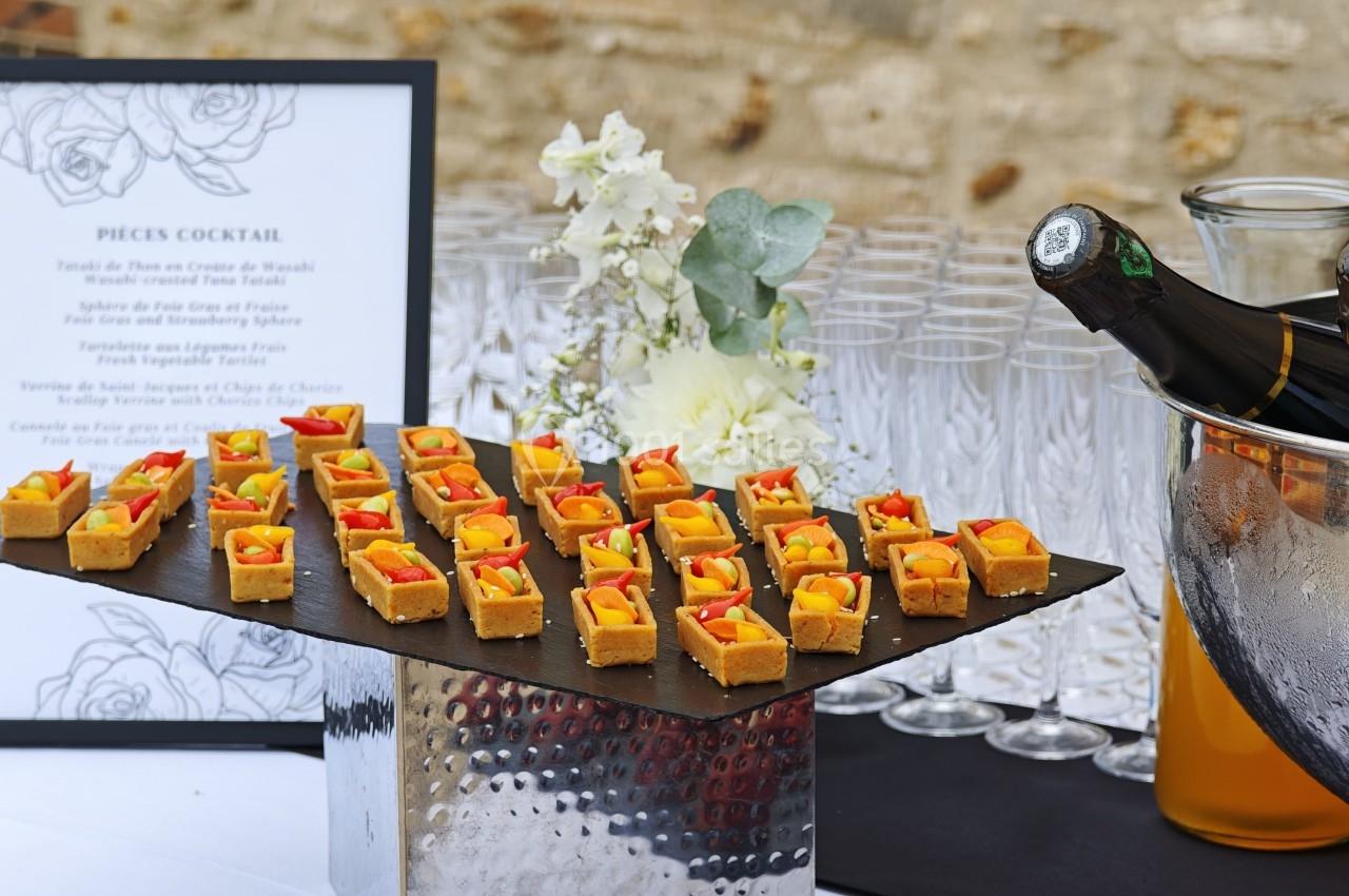 Plateau de bouchées apéritives colorées sur une table dressée avec des verres et une bouteille de champagne.
