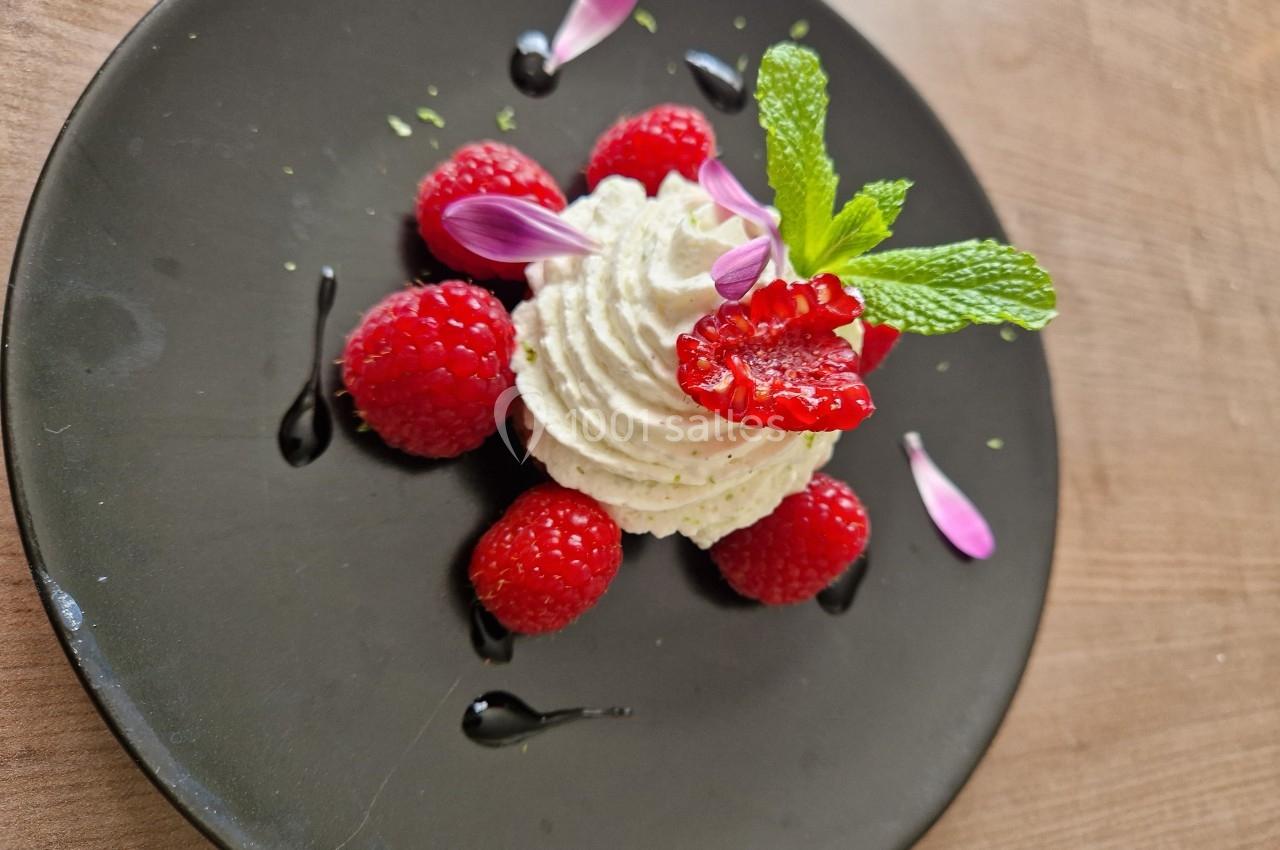 Dessert composé de crème fouettée, framboises fraîches, pétales de fleurs et feuilles de menthe sur une assiette noire.