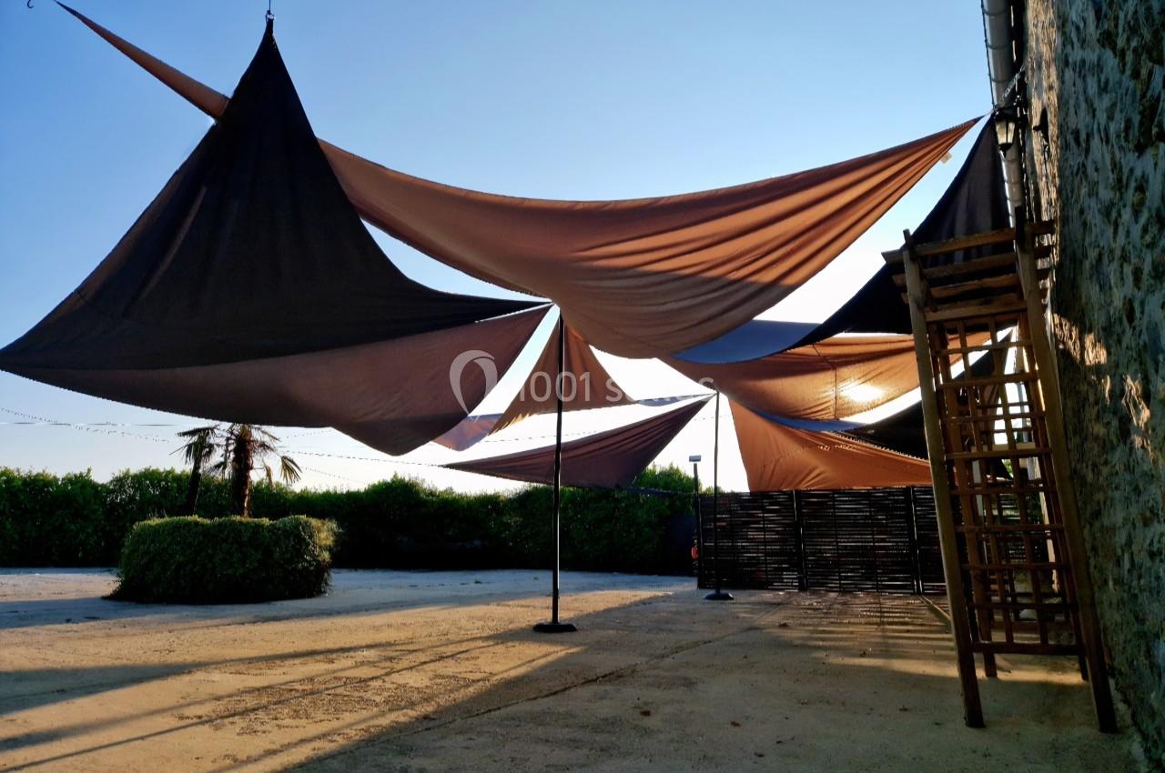 Toiles tendues créant des zones d'ombre dans une cour extérieure ensoleillée, avec un mur en pierre et des arbustes.