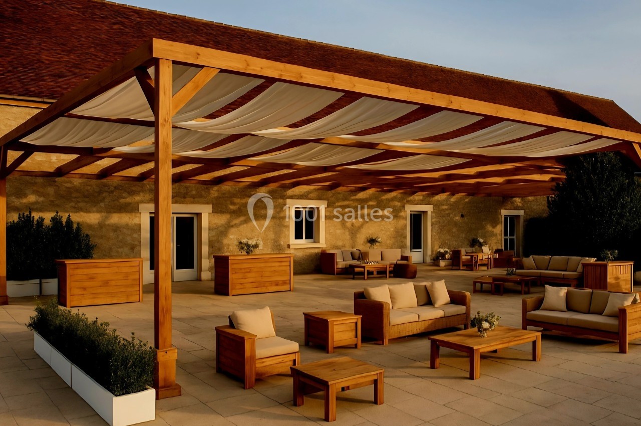 Terrasse avec pergola en bois, mobilier de jardin en bois et coussins beiges, devant un bâtiment en pierre.