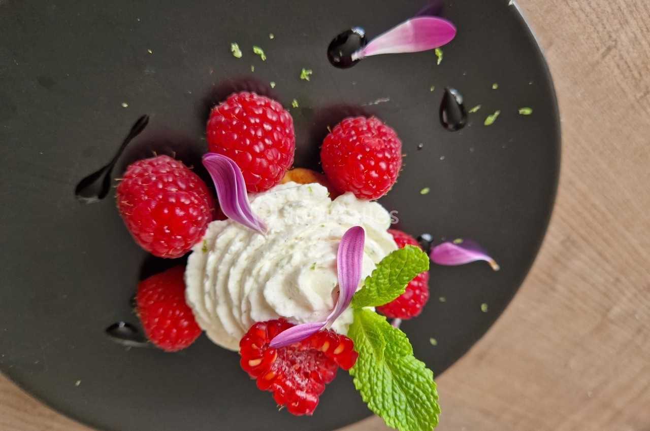 Dessert décoratif avec framboises, chantilly, pétales violets, menthe et filet de sauce sur une assiette noire.