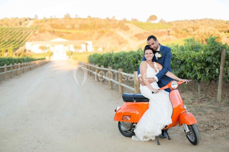 Un couple souriant en tenue de mariage assis sur un scooter orange, devant un chemin bordé de vignes.
