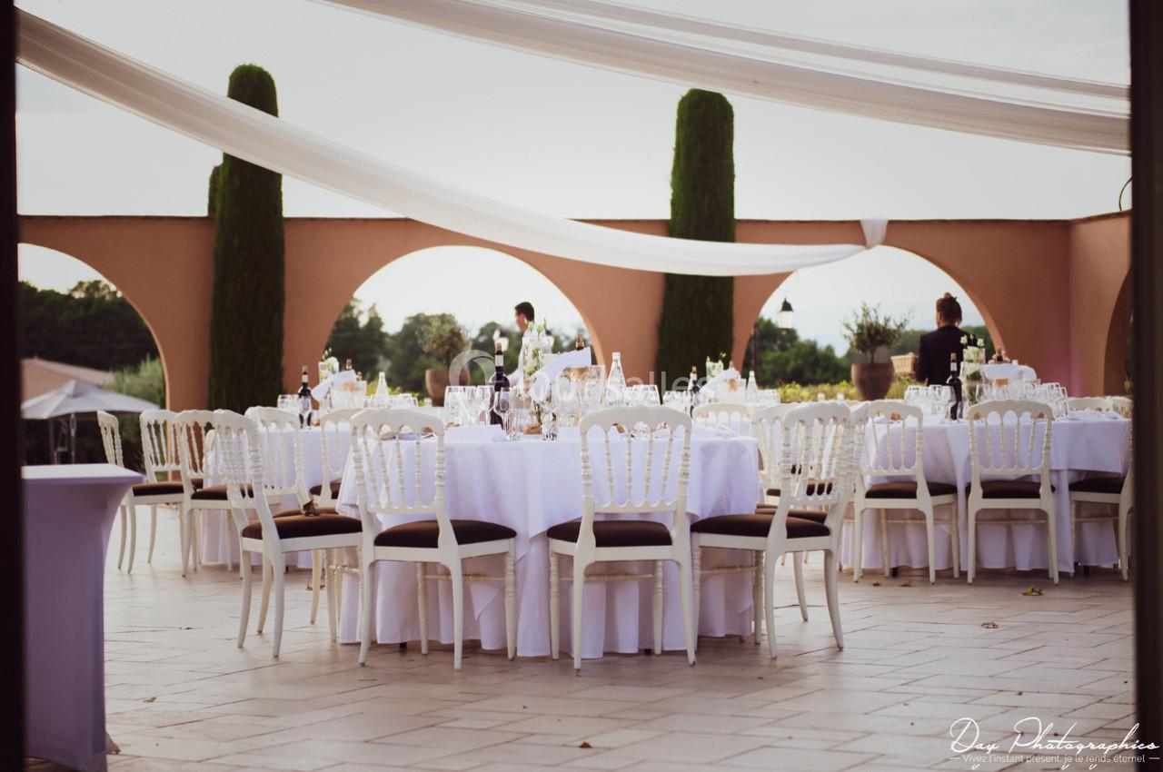 Tables rondes dressées avec nappes blanches sur une terrasse extérieure, décorée de voilages et entourée d'arches.