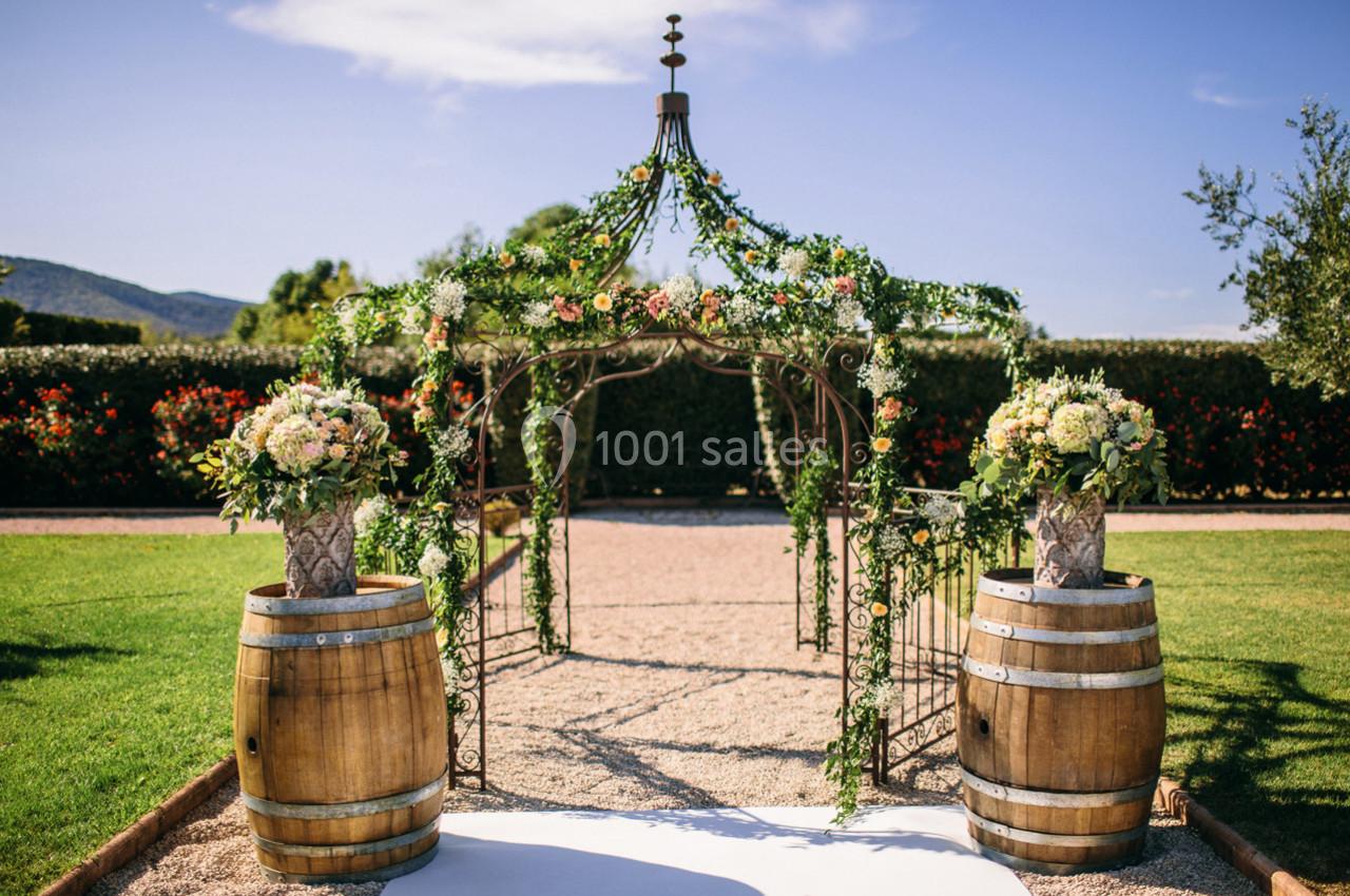 Arche de mariage décorée de fleurs et de feuillage, encadrée par deux tonneaux fleuris, dans un jardin ensoleillé.