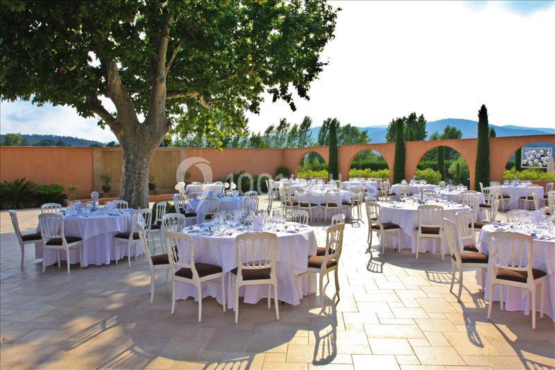 Tables dressées en extérieur sur une terrasse ensoleillée, entourée d'arches et d'un grand arbre.