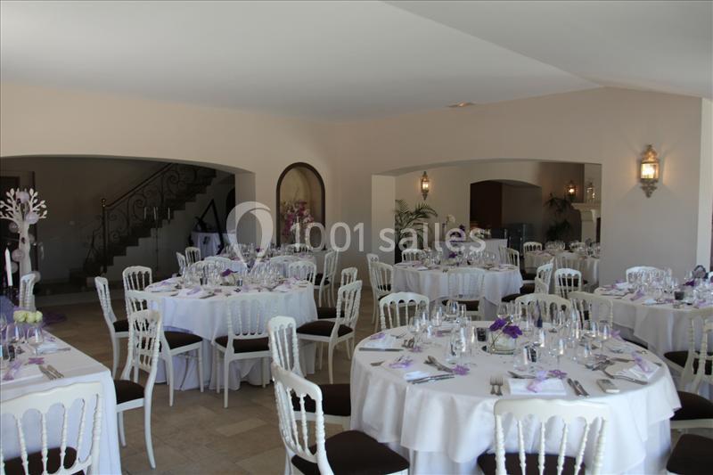 Salle de réception lumineuse avec tables rondes dressées, décorations violettes et chaises blanches.