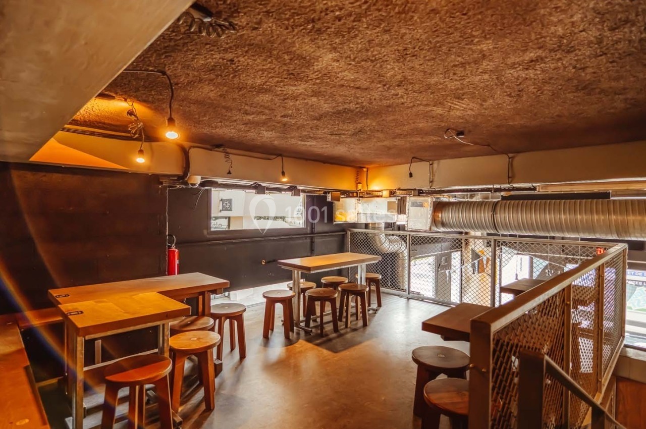 Espace intérieur avec tables en bois, tabourets et éclairage tamisé, situé à l'étage d'un lieu au style industriel.
