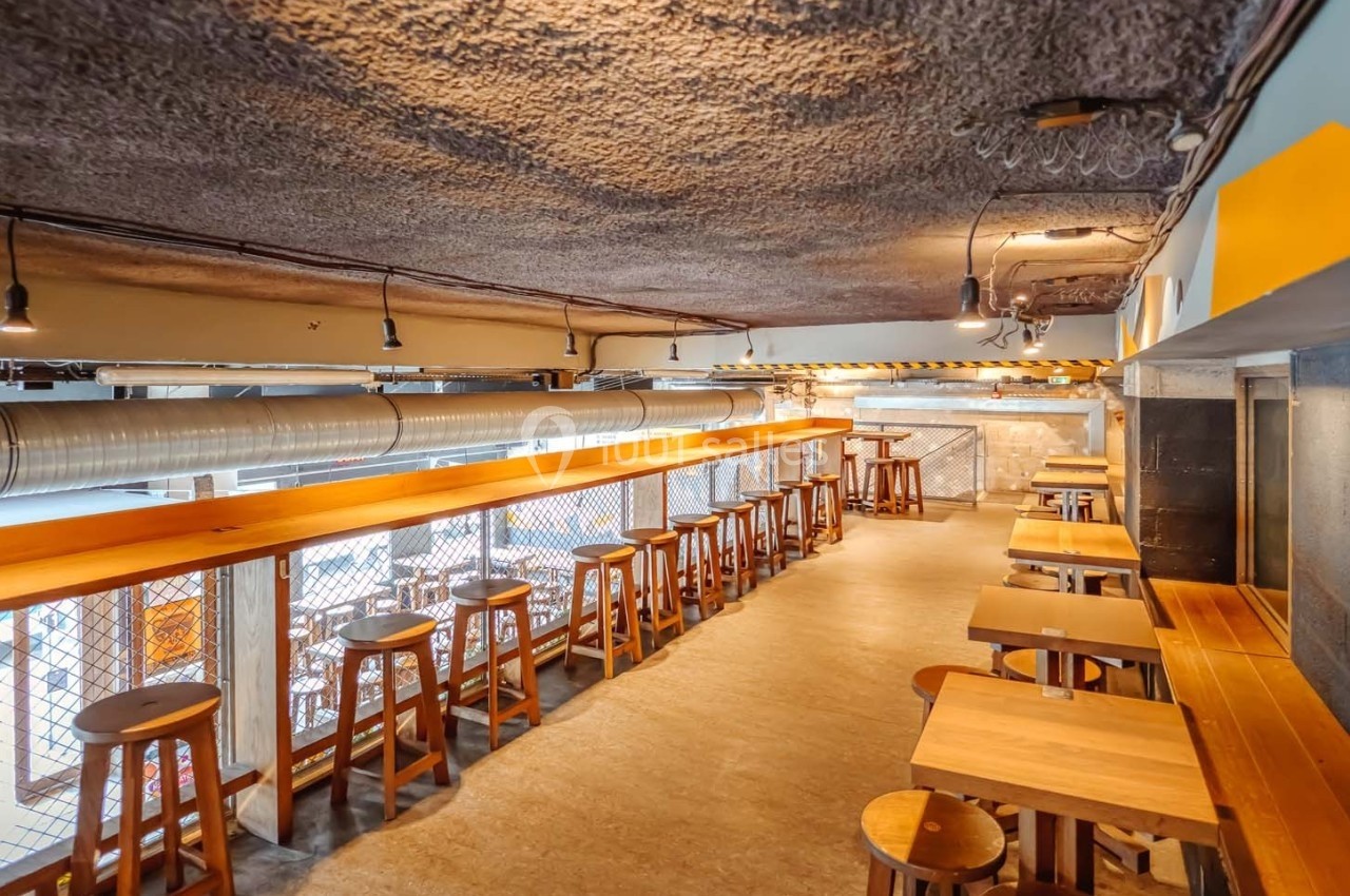 Salle avec tables et tabourets en bois, éclairage suspendu et vue sur un espace ouvert à travers une balustrade grillagée.