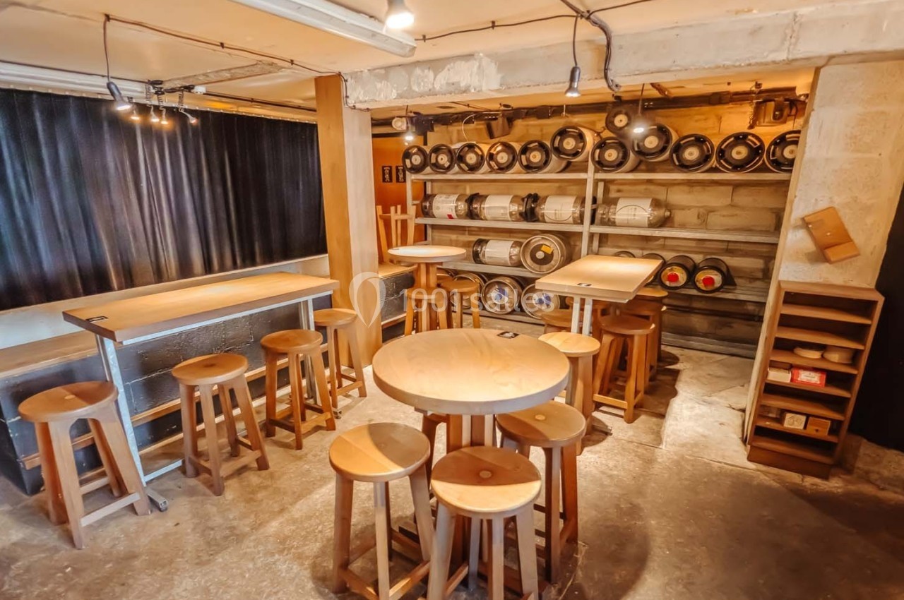 Salle avec tables et tabourets en bois, étagères contenant des fûts de bière alignés contre un mur en béton.